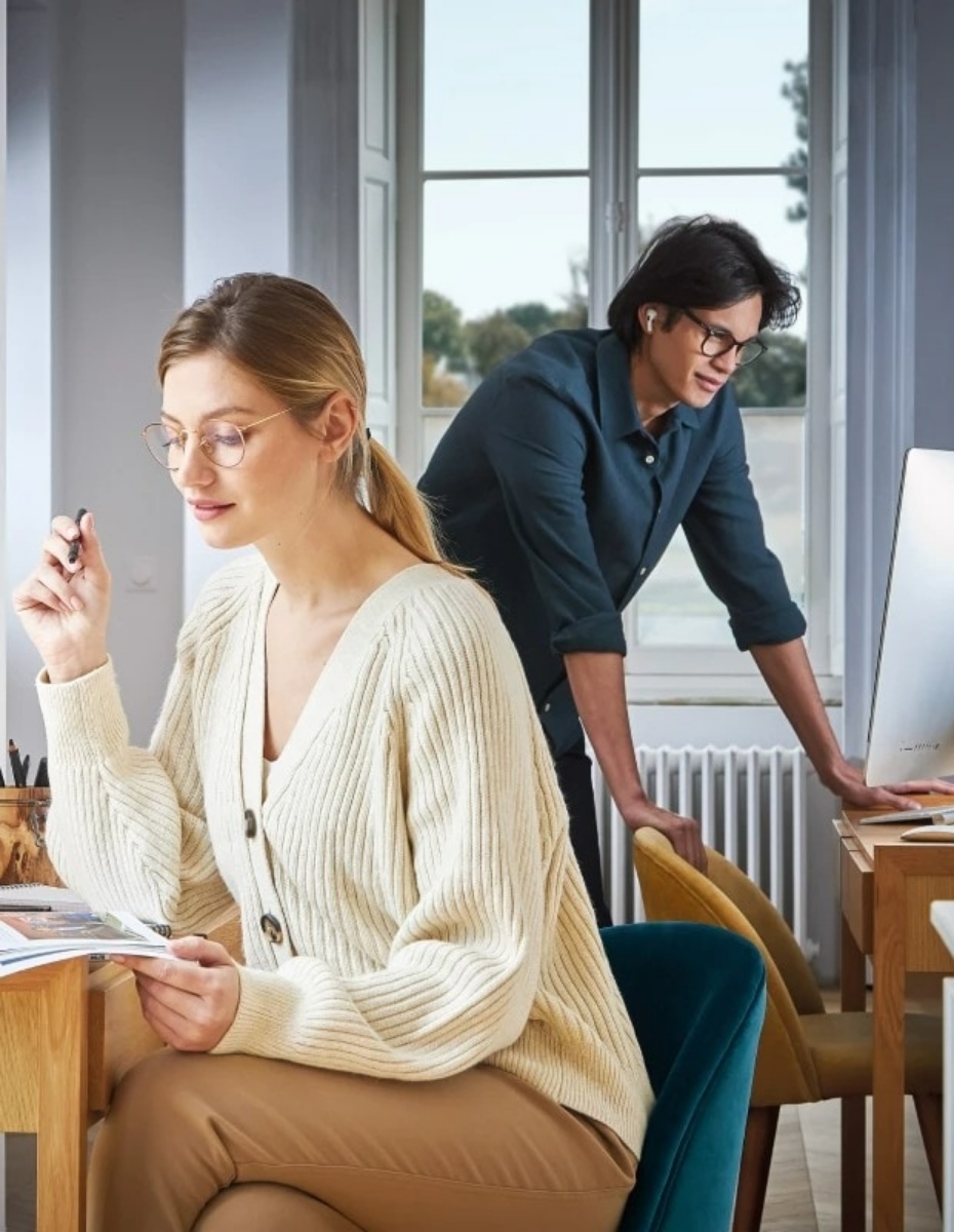 Frau mit Brille im Vordergrund liest in einem Magazin. Mann im Hintergrund mit Brille blickt in einen Bildschirm. 
